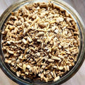 A close-up top-down view of dried, chopped herbal roots or bark pieces in a clear glass jar, showing natural shades of brown and beige.