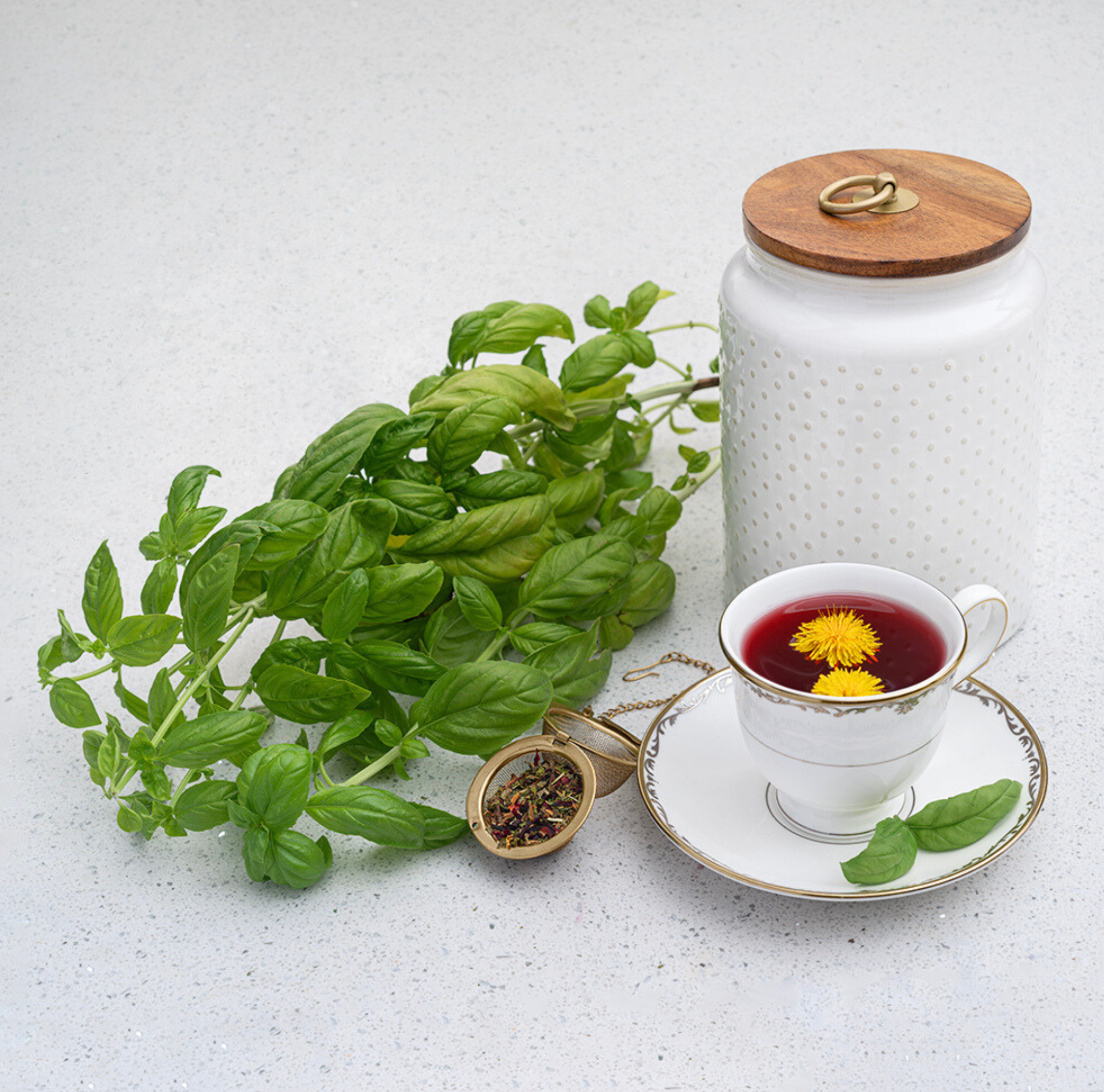 Fresh basil next to white herb container with wooden lid next to white teacup with red tea inside on a white background