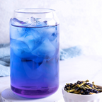 A clear glass with iced blue liquid next to a bowl of dried butterfly pea flower on a white background