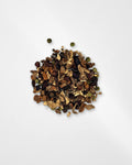 A small pile of loose dried botanical tea or spice blend with brown leaves, dark berries, and a few green round pieces, photographed on a clean white background.