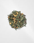 A small pile of loose dried herbal tea or botanical blend, showing green leaves, brown stems, and pale yellow dried flower pieces on a light gray background.