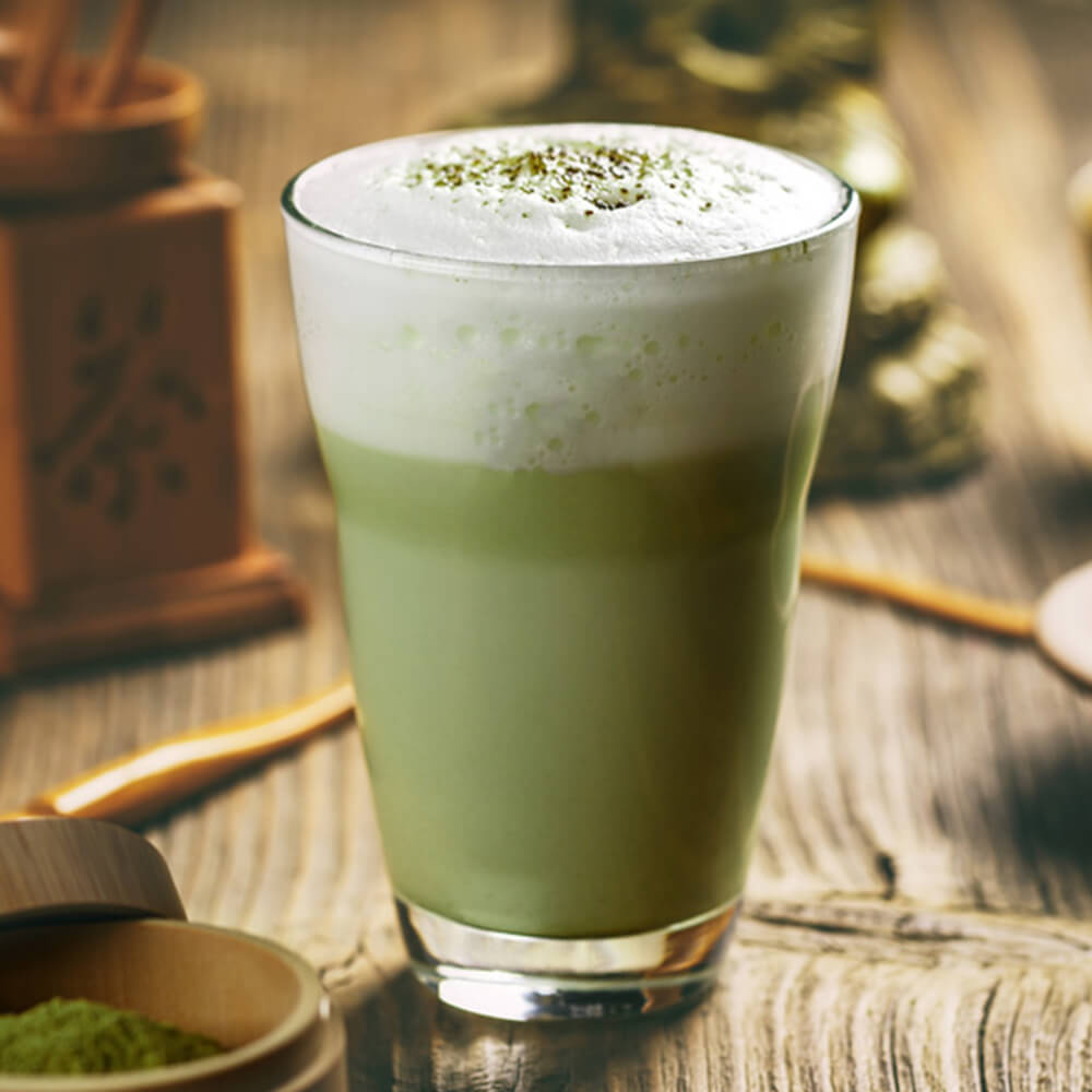 A green tea matcha in a clear glass with a frothy head sprikled with matcha powder with a blurred apothecary background