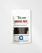 A white stand-up pouch of 'The Tea Shop' Hibiscus Mate herbal tea blend, featuring green and red text, a clear window showing the loose leaf tea inside, and a blue circular logo at the bottom, set against a light gray background.