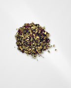 A small pile of loose-leaf herbal tea blend with dried green leaves, yellow-brown botanical pieces, and dark red accents, photographed on a clean white background.