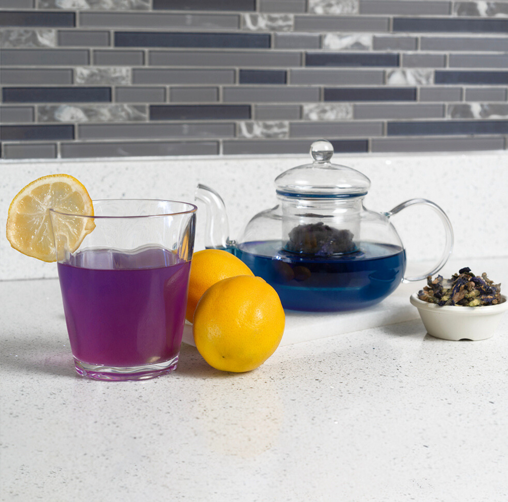 Tea set with a glass of purple liquid, lemons, and a teapot on a countertop.
