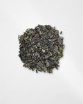 A small mound of loose-leaf tea leaves, appearing dark green to black with subtle olive and brownish highlights, photographed from above on a plain white background.