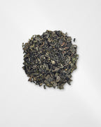 A small mound of loose-leaf tea leaves, appearing dark green to black with subtle olive and brownish highlights, photographed from above on a plain white background.