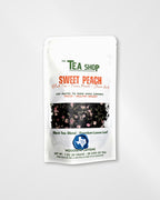 A white stand-up pouch of 'The Tea Shop' Sweet Peach tea blend, featuring green and orange text, a central photo of peach pieces, and a blue circular logo near the bottom, set against a light gray background.