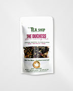A stand-up pouch of loose leaf tea from 'The Tea Shop' on a light gray background. The packaging is white with green and magenta text, featuring a clear window that displays the dark brown and black loose leaf tea blend inside.