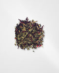 A small pile of loose-leaf herbal tea blend with dried green leaves, dark purple-red flowers, and light tan stems, photographed from above on a plain white background.