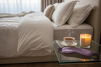 a glass nightstand with a cup of tea, a lit candle (in a jar) and a purple eye mask, next to a luxurious bed of fluffy white bedding