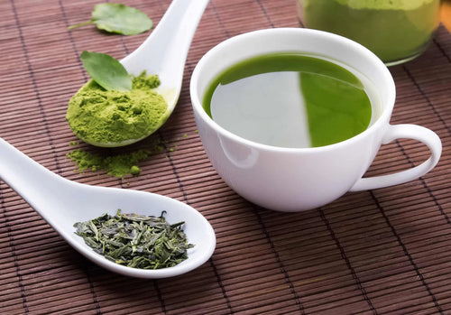 A split image showing green tea leaves on the left and Green tea matcha powder just above next to a white mug of prepared matcha on a wood colored surface