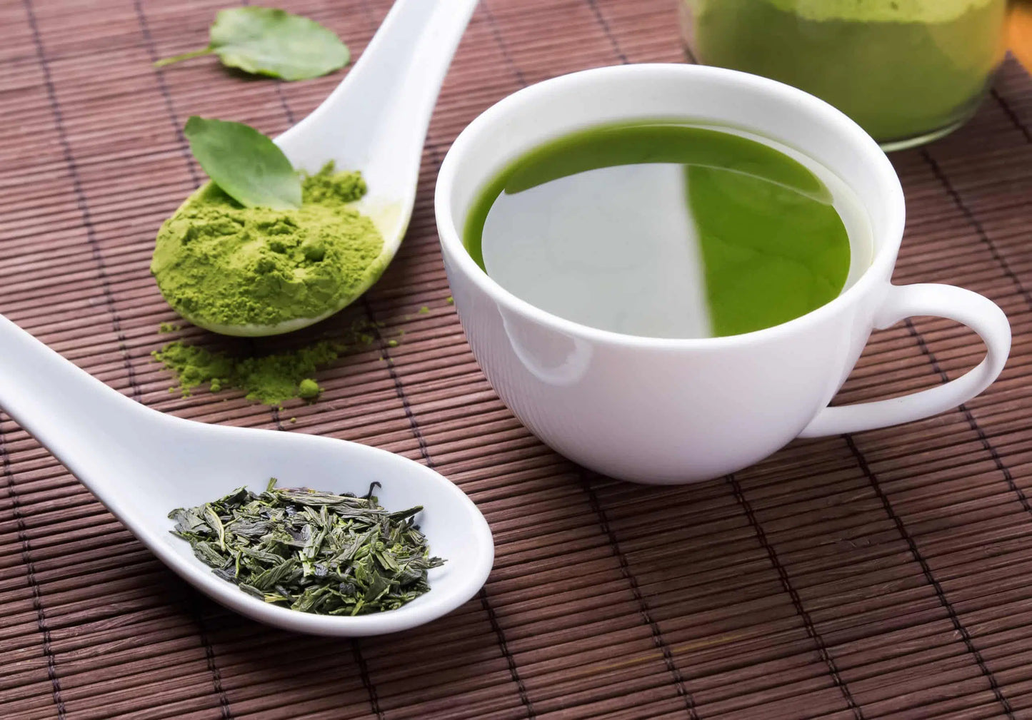 A split image showing green tea leaves on the left and Green tea matcha powder just above next to a white mug of prepared matcha on a wood colored surface