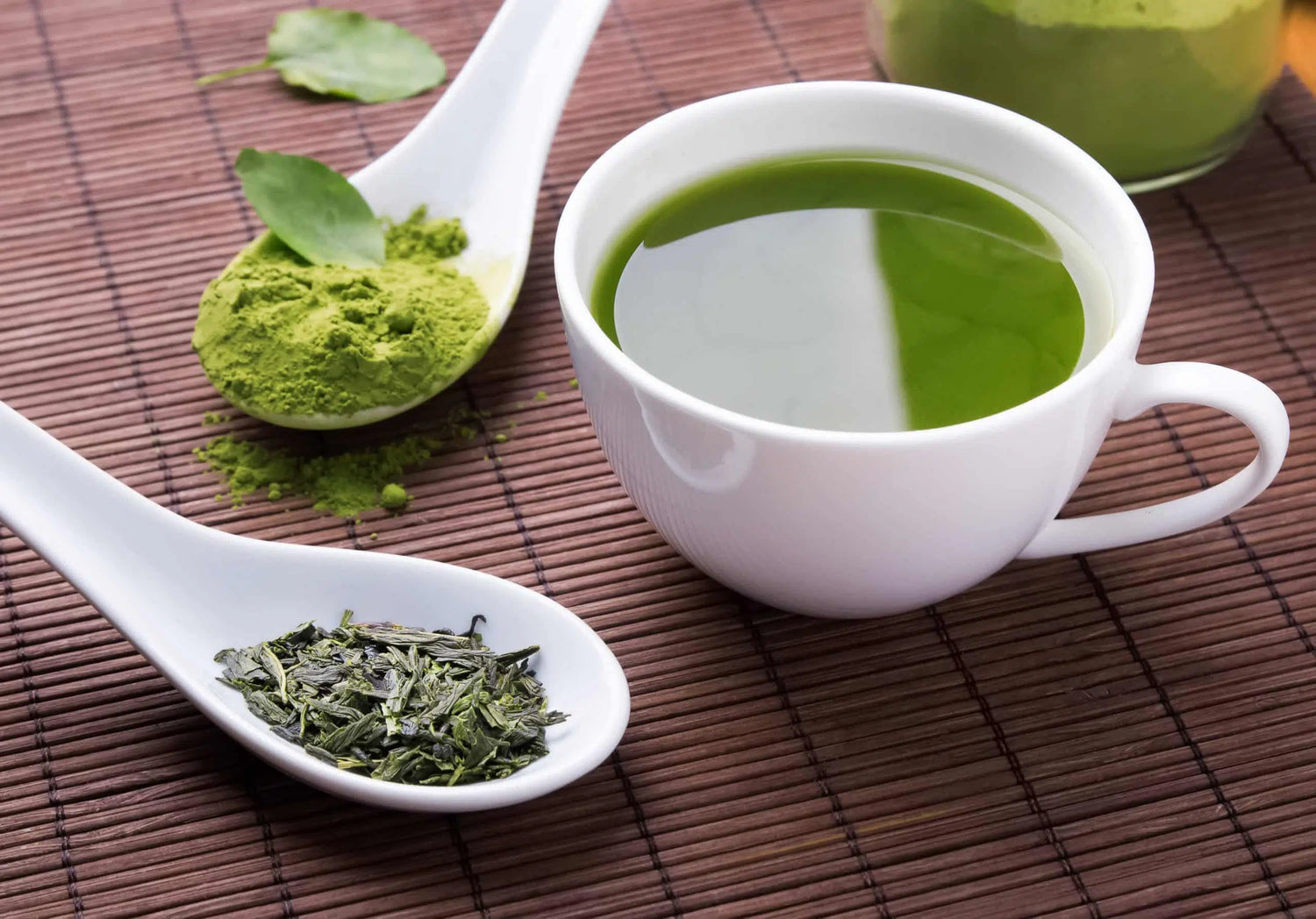 A split image showing green tea leaves on the left and Green tea matcha powder just above next to a white mug of prepared matcha on a wood colored surface